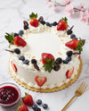 A round white frosted cake decorated with fresh strawberries, blueberries, blackberries, whipped cream, and chocolate shavings, served on a gold cake board with a small bowl of berry sauce and a gold fork on a light marble surface.