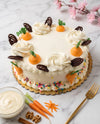 A frosted round cake decorated with piped white swirls, orange carrot toppers with green leaves, and dark chocolate curls, set on a marble surface with a gold fork, a bowl of cream, and small carrot props.