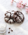 Three chocolate crinkle cookies dusted with powdered sugar, arranged on a white marble surface with a small glass of powdered sugar, scattered cocoa powder, and pink blossoms in the background.