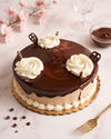 Chocolate cake with glossy chocolate glaze, piped cream rosettes, chocolate cookie pieces, and a gold cake board, styled on a light marble surface with pink blossoms and a small bowl of chocolate sauce in the background.