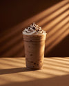 A tall clear plastic cup filled with a light brown iced coffee or milkshake, topped with whipped cream and chocolate drizzle, set on a brown surface with warm sunlight and shadow streaks.