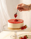 A strawberry cheesecake on a glass cake stand, topped with fresh strawberry jam, with a hand holding a spoon dripping jam above the cake and strawberries on the side.