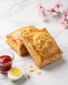 Two golden-brown flaky puff pastry boxes arranged on a white marble surface, with a small glass of red sauce, a halved hard-boiled egg, and scattered pepper flakes nearby. Pink blossoms appear softly blurred in the upper right.