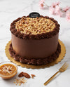 A chocolate cake with caramel frosting and pecan topping, displayed on a gold scalloped cake board with a small bowl of caramel sauce, a gold fork, and a La Crème Bakery Cafe sign on a white marble surface.