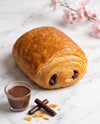 A golden-brown chocolate croissant pastry on a white marble surface, served with a small glass of chocolate sauce, chocolate sticks, and pink blossoms in the background.