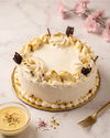 A round white frosted cake with piped cream borders, gold crumb dusting, and dark chocolate decorations, served on a gold-rimmed plate on a light marble surface with a small bowl of yellow sauce, a gold fork, and pink blossoms in the background.