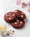 Three red velvet chocolate chip cookies with white chocolate chips are arranged on a white marble surface, with a small glass bowl of white chocolate chips and pink blossoms in the background.
