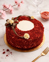 A red velvet cake with a white cream center and chocolate ribbon decorations, presented on a gold cake board on a white marble surface with a gold fork, pink blossoms, and a glass in the background.