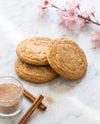 Three golden-brown sugar cookies stacked on a white marble surface, styled with a small glass jar of cinnamon sugar, cinnamon sticks, and pink cherry blossoms.