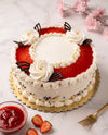 A round strawberry cake with white frosting, piped cream roses, a red strawberry topping, and chocolate decorations, presented on a gold scalloped cake board with a small bowl of strawberry sauce, sliced strawberries, and a gold fork on a light marble surface.