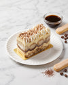 A slice of layered tiramisu cake with whipped cream and cocoa powder topping, served on a white paper plate with a gold base. A small glass bowl of espresso, ladyfinger cookies, and coffee beans are arranged on a white marble surface.