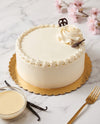 A round white frosted cake with piped border decoration, a cream rose topper, and chocolate garnish, displayed on a gold scalloped cake board on a marble surface with a small bowl of frosting, vanilla beans, and a gold fork nearby.