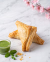 Two golden-brown flaky pastry triangles stacked on a white marble surface, with a small glass bowl of green chutney, mint leaves, and a few green peas nearby. Pink blossoms are softly blurred in the background.