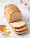 A loaf of sliced whole wheat bread on a white marble surface, styled with a small glass of honey, a butter cube, wheat stalks, and pink blossoms in the background.
