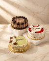 Three decorated birthday cakes displayed on white pedestals against a beige marble background. The cake on the left is chocolate with chocolate decorations and a green center; the middle cake is white with red fruit and white cream; the cake in front is light green with white cream and chocolate drizzle.