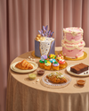 A styled dessert table draped in a beige tablecloth, featuring a pink tiered cake, a lavender cake with gold and purple butterfly decorations, colorful cupcakes, a croissant, a brownie, and small dessert dips against a soft pink curtain backdrop.